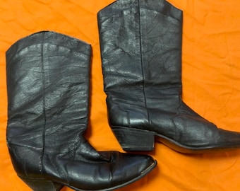 Op de afbeelding: Een paar zwarte leren cowboylaarzen. De laarzen hebben een puntige neus, een hak van gemiddelde hoogte en een hoge schacht. Het leer lijkt versleten en getextureerd. De laarzen staan tegen een oranje achtergrond.