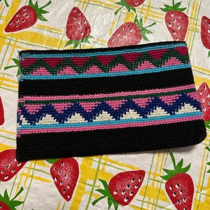 May include: A black, rectangular clutch with a colorful, geometric pattern. The pattern includes pink, blue, and white zigzags and stripes. The clutch is set against a background of a yellow and white tablecloth with red strawberries.