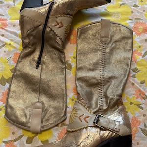 Puede incluir: Un par de botas doradas hasta la rodilla con un acabado met&aacute;lico envejecido. Las botas tienen punta afilada, tac&oacute;n ancho y costuras decorativas. Una bota tiene cremallera lateral, la otra hebilla. Detalles en cuero beige y dise&ntilde;o de sol.