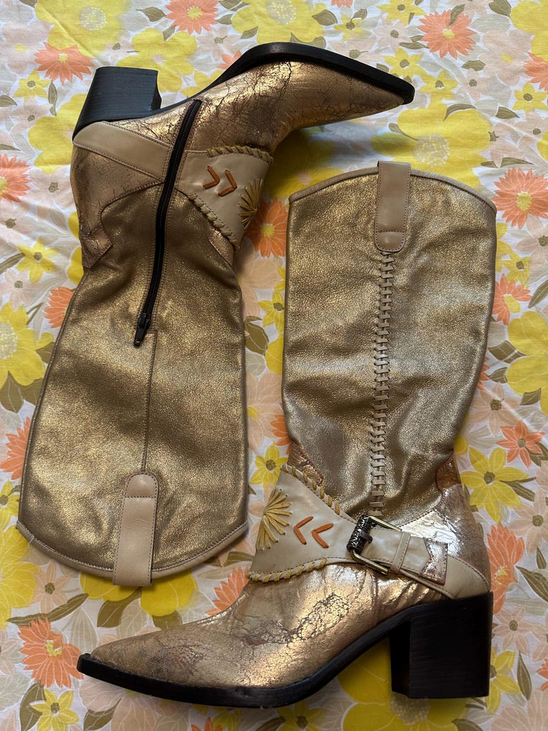 Puede incluir: Un par de botas doradas hasta la rodilla con un acabado met&aacute;lico envejecido. Las botas tienen punta afilada, tac&oacute;n ancho y costuras decorativas. Una bota tiene cremallera lateral, la otra hebilla. Detalles en cuero beige y dise&ntilde;o de sol.