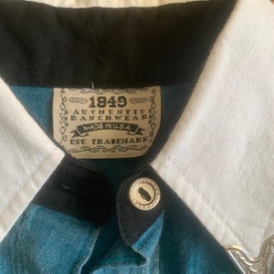May include: Close-up of a denim shirt with a white collar and black trim. The shirt features silver buttons and a decorative silver collar tip. The label reads "1849 Authentic Ranchwear Made in USA".