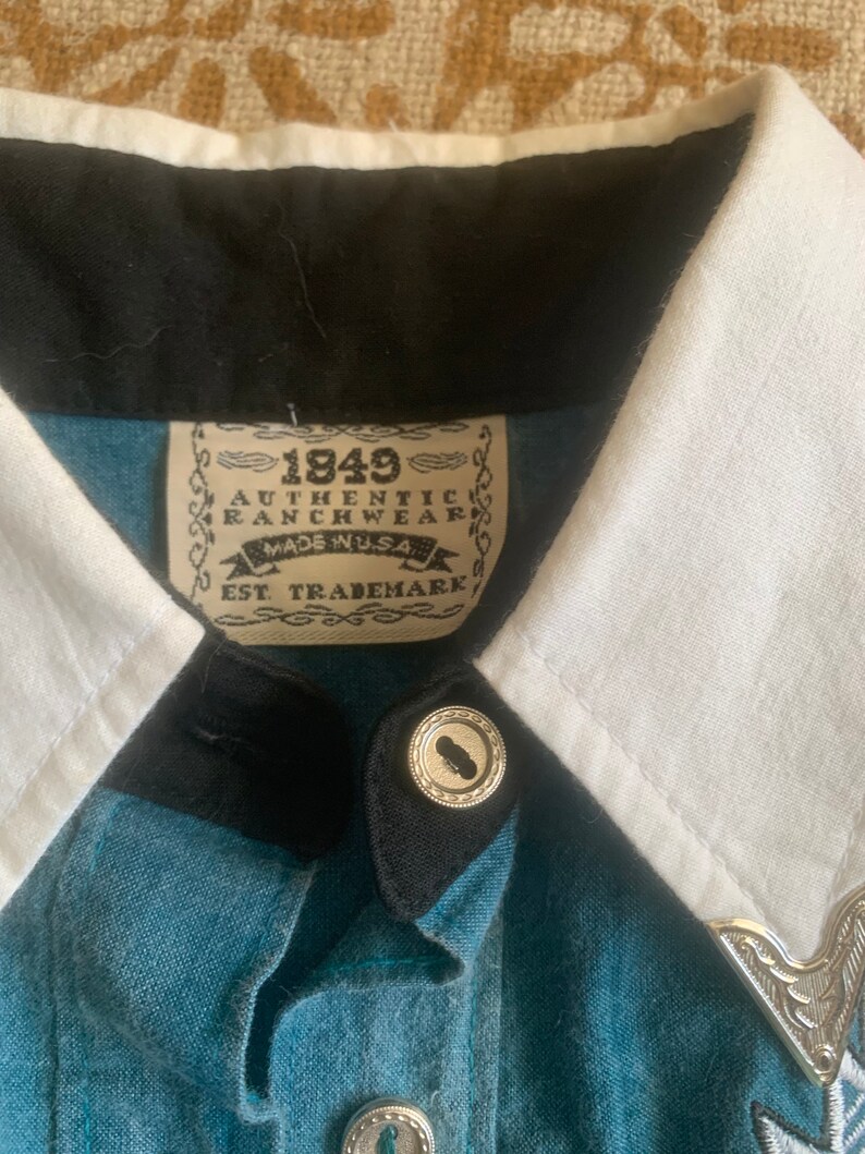 May include: Close-up of a denim shirt with a white collar and black trim. The shirt features silver buttons and a decorative silver collar tip. The label reads "1849 Authentic Ranchwear Made in USA".
