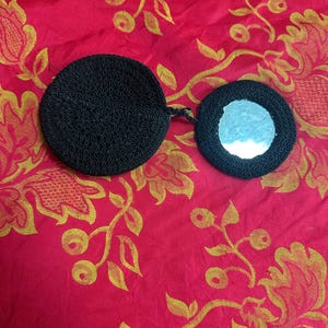 May include: A black, round, vintage compact mirror with a matching cover, connected by a short strap. The mirror is surrounded by a black, textured border. The compact is set against a red background with a gold floral pattern.