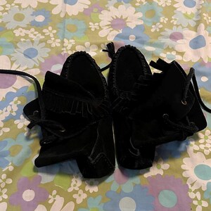 May include: A pair of black suede moccasins with fringe detailing and laces. The moccasins are on a floral patterned fabric background.