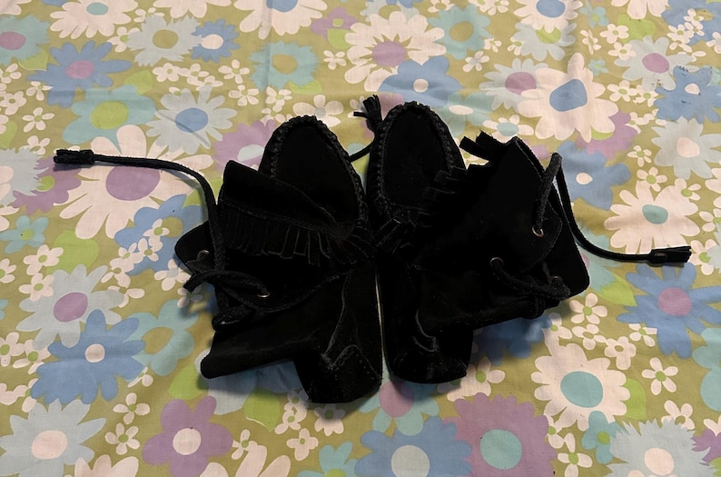 May include: A pair of black suede moccasins with fringe detailing and laces. The moccasins are on a floral patterned fabric background.