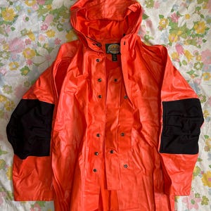 May include: Bright orange hooded rain jacket with black patches on the shoulders. The jacket has a double-breasted closure with multiple buttons and a label on the chest. The jacket is made of a shiny, waterproof material.