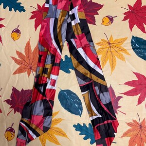 May include: A long, narrow silk scarf featuring a geometric pattern in shades of red, black, white, and gold. The scarf has a smooth, reflective finish and is arranged in an inverted V-shape, showcasing a repeating abstract design of blocks and lines.