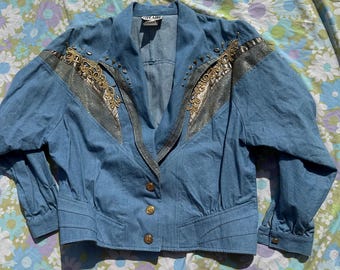 May include: Light blue denim jacket with gold-colored embellishments on the shoulders and upper chest. The jacket has a V-neck collar, long sleeves, and gold buttons. The jacket has a vintage style.