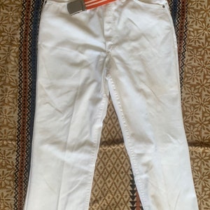 Puede incluir: Pantalón blanco de pierna ancha con un cinturón ancho de rayas naranja y blancas. El cinturón tiene una hebilla plateada.