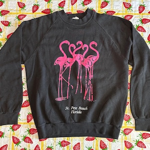 May include: A dark gray sweatshirt featuring a pink flamingo graphic. The graphic includes three flamingos and the text "St. Pete Beach Florida". The sweatshirt has long sleeves and a crew neck.