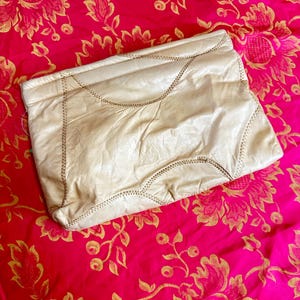 Peut inclure: Une pochette en cuir crème avec des détails cousus. Le sac rectangulaire a un design souple et plié et est posé sur un fond floral rose vif. Les coutures sont visibles le long des bords et des coutures de la pochette.