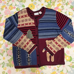 May include: A patchwork cardigan sweater in shades of blue, red, and tan. The sweater features a Christmas tree and a teddy bear applique. The sleeves and front panels have geometric patterns and stripes. The sweater has a button-up front.