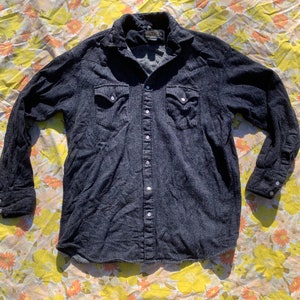 May include: A dark blue denim long-sleeved shirt with a button-down front and two chest pockets. The shirt has a slightly faded look and a textured fabric.