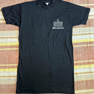 May include: Black t-shirt with short sleeves. The shirt features a small graphic on the front with the text "Cactus Saloon Daytona Beach, Fla. Bike week 82". The shirt is laid flat on a striped fabric.