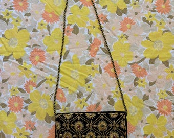 May include: Black rectangular shoulder bag with gold and jewel embellishments. The bag features a floral pattern with gold thread and red and green jewels. The strap is a twisted gold and black cord. The background is a floral pattern in yellow, orange, and white.