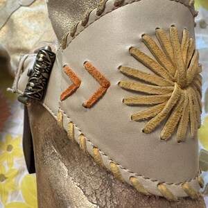Puede incluir: Primer plano de una bota dorada con un brazalete de cuero color crema. El brazalete presenta costuras en forma de flecha naranja y un dise&ntilde;o de sol. La bota tiene un cierre met&aacute;lico decorativo y detalles cosidos.