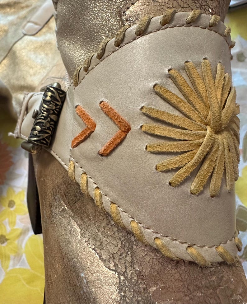 Puede incluir: Primer plano de una bota dorada con un brazalete de cuero color crema. El brazalete presenta costuras en forma de flecha naranja y un dise&ntilde;o de sol. La bota tiene un cierre met&aacute;lico decorativo y detalles cosidos.