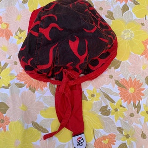 May include: A red and black head wrap with a flame design. The head wrap has a red tie and a small white tag with a Native American graphic. The head wrap is made of fabric.