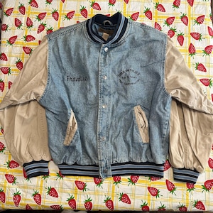 May include: A blue denim varsity jacket with tan sleeves and black and blue striped ribbing. The jacket has a button closure and two pockets. The text "Frankie" is embroidered on the left side of the jacket and "Brewskies Pub & Eatery The Southeastern" is embroidered on the right side.