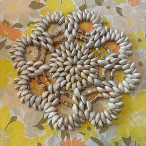 Puede incluir: Un posavasos circular hecho a mano con pequeñas conchas marinas de color crema. El posavasos presenta un diseño floral con las conchas dispuestas en un patrón repetitivo. Un artículo decorativo para el hogar.