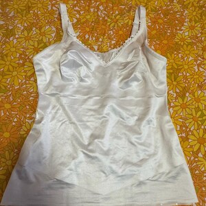 Könnte beinhalten: Ein weißes Satin-Camisole mit verstellbaren Trägern und einem V-Ausschnitt. Das Oberteil hat eine zarte Spitzenborte am Ausschnitt und eine glatte, seidige Textur. Das Kleidungsstück ist auf Komfort und Stil ausgelegt.