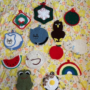 Könnte beinhalten: Eine Sammlung von 12 gehäkelten Topflappen in verschiedenen Formen und Farben, darunter eine Katze, ein Frosch, eine Wassermelone, ein Regenbogen, ein Apfel, eine Schneeflocke, ein Kranz, eine Katze, eine Sonne, ein Vogel und ein Gesicht.
