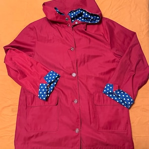May include: A red raincoat with a hood and button closure. The interior of the hood and the cuffs are lined with a blue fabric featuring white polka dots. The jacket has two front pockets.