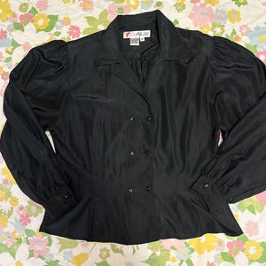 May include: Black double-breasted blouse with long sleeves and a collared neckline. The blouse features a fitted waist and puffed sleeves. The fabric has a slight sheen. The blouse has a label with the text "CALS".