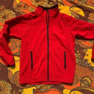 Puede incluir: Una chaqueta softshell roja con cremallera negra y ribetes negros. La chaqueta tiene dos bolsillos con cremallera en la parte delantera.