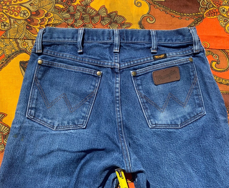 Puede incluir: Un par de pantalones vaqueros azules de mezclilla con un parche de cuero marr&oacute;n en el bolsillo trasero que dice "Wrangler".