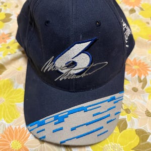 Puede incluir: Una gorra de béisbol azul marino con un diseño bordado en blanco y azul. El número "6" se muestra de forma prominente en la parte delantera de la gorra, junto con una firma.