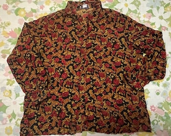 Op de afbeelding: Een overhemd met lange mouwen en knoopsluiting op een zwarte ondergrond, voorzien van een dicht, herhalend patroon van rode bloemen en goudkleurige paisleyvormen. Het kledingstuk is afgewerkt met een kraag en knoopmanchetten.