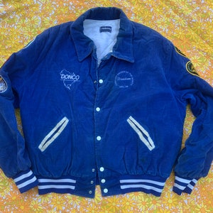 Puede incluir: Chaqueta de béisbol de pana azul con ribetes de rayas blancas y azules. La chaqueta tiene cierre de botones y presenta parches bordados con el texto "Donco" y "University of Davidson".