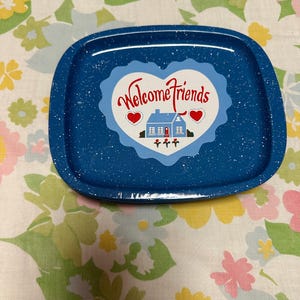 Puede incluir: Una pila de bandejas de esmalte azul con motas blancas. Cada bandeja tiene un gráfico en forma de corazón con las palabras "Welcome Friends", una casa y corazones rojos. Las bandejas están sobre un mantel con estampado floral.