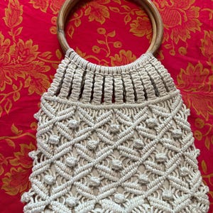 以下が含まれることがあります： クリーム色のマクラメハンドバッグ。幾何学模様と木製の持ち手が付いています。バッグはダイヤモンド型と小さなテクスチャードノットの織り模様のデザインです。持ち手は滑らかなライトブラウンの木材でできており、クリーム色のバッグとの自然なコントラストを生み出しています。