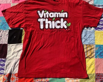 Puede incluir: Camiseta roja con un gráfico blanco que dice "Vitamin Thick" con una hoja verde y una cereza roja. El texto debajo del gráfico dice "It was all a dream."