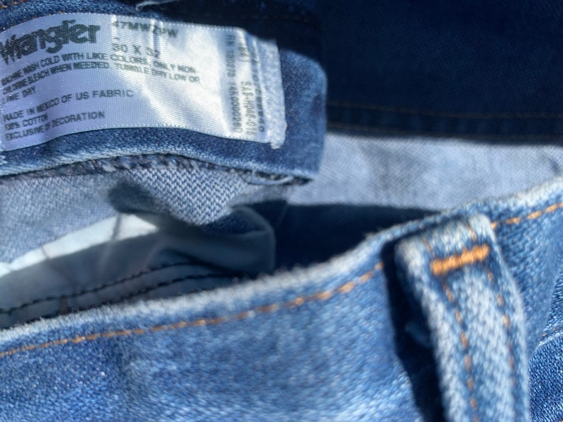 Puede incluir: Primer plano de una etiqueta de mezclilla azul con texto blanco que dice "Wrangler 47MW 4PW 30 X 32", "Lavar a m&aacute;quina con agua fr&iacute;a con colores similares, solo blanqueador sin cloro cuando sea necesario, secar en secadora a baja temperatura, secar en l&iacute;nea", "Hecho en M&eacute;xico con tela de EE. UU., 100 % algod&oacute;n, exclusivo de la decoraci&oacute;n".