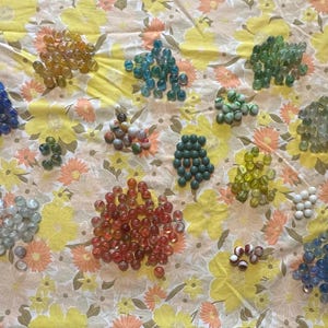 Puede incluir: Una colección de canicas de vidrio de colores en varios tamaños y colores, incluyendo azul, verde, rojo y amarillo. Las canicas están dispuestas sobre una tela amarilla con estampado floral.