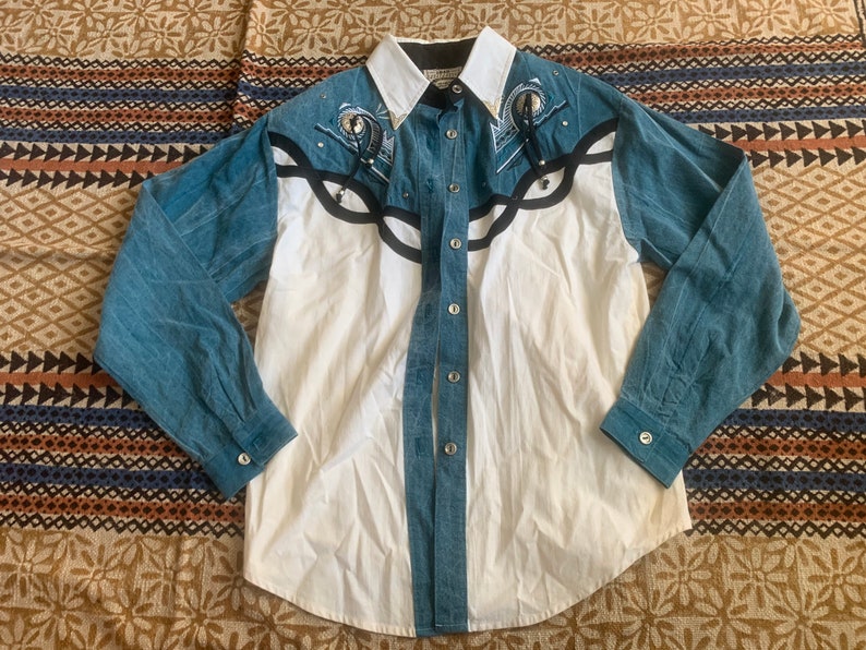 May include: A button-down shirt with a white body, blue denim sleeves, and a white collar. The shirt features black and silver decorative accents on the front and sleeves. The shirt has a Western style.