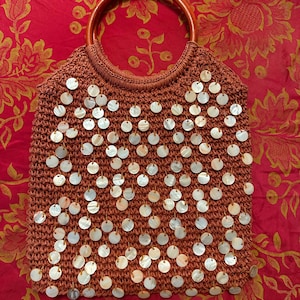 以下が含まれることがあります： 茶色の編みバッグ、丸い木製の持ち手付き。 バッグには、多数の白く円形の装飾が施されています。 バッグは、花柄の赤い背景に置かれています。