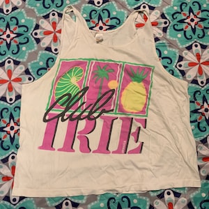 May include: White tank top with a pink and green graphic design. The graphic features a pineapple, a palm tree, and a seashell. The text "Club Tribe Jamaica" is printed on the graphic.