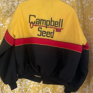 Puede incluir: Una chaqueta amarilla, negra y roja con cierre de cremallera. La parte posterior de la chaqueta tiene un logotipo que dice "Campbell Seed" con un gráfico de llave.