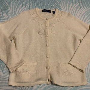 May include: Cream-colored cardigan sweater with floral embroidery. The button-down sweater has two front pockets and a scalloped hem. The brand name "Karen Scott" is visible on the label. The sweater is made of a soft, textured knit fabric.