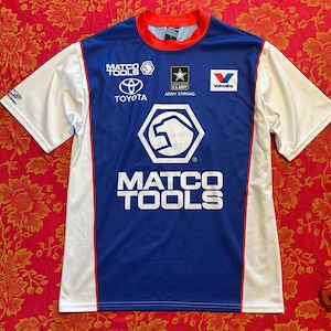 Può includere: Una maglia blu e bianca a maniche corte con finiture rosse e colletto rosso. La maglia presenta il logo "MATCO TOOLS", il logo Toyota, il logo dell'esercito americano e il logo Valvoline.
