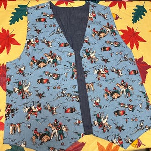 May include: A sleeveless vest featuring a light blue fabric with a repeating pattern of figures on horseback and bison. The interior lining is a solid dark blue fabric. The vest has a V-neckline and a pointed hemline at the front opening.