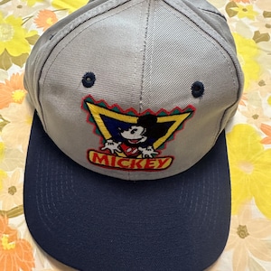Pode incluir: Boné de beisebol cinza com aba azul marinho. O boné apresenta um design bordado do Mickey Mouse em vermelho, amarelo, azul e preto, com a palavra "MICKEY" abaixo. Possui um pequeno botão azul marinho no topo.
