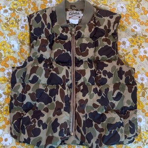 Puede incluir: Chaleco acolchado de camuflaje de la marca Cabela's con cierre de cremallera. El chaleco está hecho de un tejido acolchado y tiene un esquema de color marrón, verde y negro.