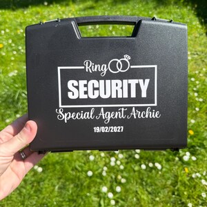 May include: Black case with a handle, featuring the text "Ring Security" and "Special Agent Archie 19/02/2027" in white. The case has a wedding ring graphic above the word "Security".