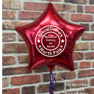 Op de afbeelding: Een glanzende rode stervormige ballon met de tekst "Special Delivery" en "Special elf Delivery for Wynter North Pole" in een witte cirkel. De ballon heeft een blauw en rood lint. Een "Elf Delivery Sticker" is ook zichtbaar.