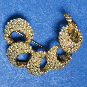 May include: A gold-toned brooch with a design of multiple loops of faux pearls. The brooch is in the shape of a crescent moon.
